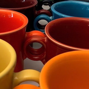 May include: A collection of colorful ceramic mugs with handles. The mugs are stacked in a pyramid shape, with the largest mug at the bottom and the smallest mug at the top. The mugs are in various shades of red, orange, yellow, green, blue, and brown.
