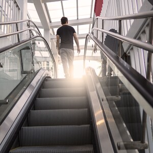 May include: A person walks up a glass and metal escalator with a white ceiling and red and white striped walls.