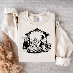 May include: A cream-colored sweatshirt with a black and white illustration of the nativity scene. The illustration depicts Mary, Joseph, and baby Jesus in a manger with two animals.