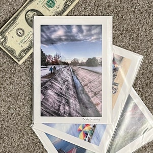 May include: Four white photo cards with different images. The top card shows a view of a concrete channel with two people walking on a path to the left. The sky is cloudy with a blue hue. The text "Brisk Serenity" is printed on the bottom of the card. The second card shows a colorful hot air balloon with a blue sky and white clouds. The third card shows a view of a mountain range with a red building in the foreground. The fourth card shows a view of a beach with a blue sky and white clouds.