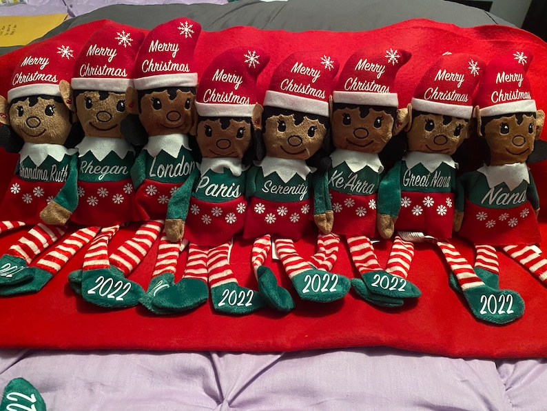 May include: Seven Christmas elf dolls with dark skin tones, wearing red hats with "Merry Christmas" and white snowflakes. Each elf has a name embroidered on its green shirt. Their legs are red and white striped with "2022" on the feet.