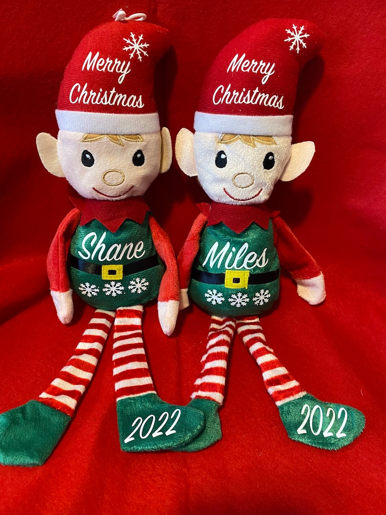 May include: Two Christmas elf soft toys. Each has a red hat with "Merry Christmas" embroidered, a green tunic with a name and snowflakes, and red and white striped legs. The year "2022" is embroidered on the green feet.