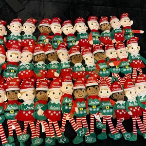 May include: A group of Christmas elf soft toys, each with a name embroidered on their green or red outfits. The elves wear red hats with "Merry Christmas" and snowflakes. The year "2021" is embroidered on their feet.