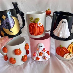 Puede incluir: Seis tazas de cerámica con decoraciones de temática de Halloween. Las tazas presentan calabazas, fantasmas y un gato negro. Las tazas son de diferentes colores, incluyendo blanco, negro, rosa y dorado.