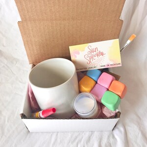 May include: A white ceramic mug, a small jar of white paint, six colorful paint cubes, a red glue stick, a paintbrush, and a business card with the text "Sun Sprinkles Handcrafted Jewelry Made in Michigan".