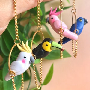 May include: Three colorful bird charms on gold chains. The birds are a pink cockatoo, a black toucan, and a blue parrot. Each bird is attached to a gold hoop.