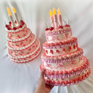 May include: Two pink and white tiered cakes decorated with cherries and white frosting. The cakes have candles on top and are on a white surface.