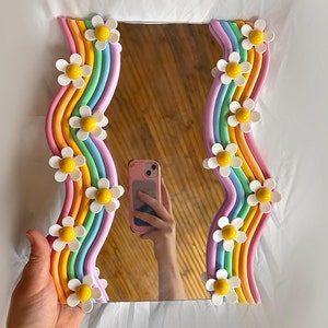 May include: A rectangular mirror with a wavy rainbow-colored frame. The frame is decorated with white daisy-shaped flowers with yellow centers. The mirror is being held up by a hand.
