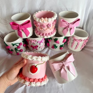 Puede incluir: Una colección de ocho macetas de cerámica rosa y blanca decoradas con diversos adornos, incluyendo lazos, cerezas, fresas y glaseado.
