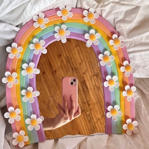 May include: A rainbow-coloured mirror with a daisy design. The mirror is shaped like a rainbow arch and has a white background. The daisies are in various shades of pink, yellow, and white.
