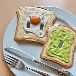 May include: A white plate with two decorative light switch covers in the shape of toast. One is topped with a fried egg, the other with green avocado. A silver fork and knife are also on the plate.