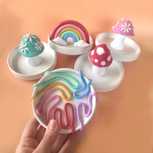 Puede incluir: Cuatro peque&ntilde;os platos de cer&aacute;mica blanca con decoraciones de arcilla de colores. Cada plato presenta un dise&ntilde;o diferente: un arco&iacute;ris, un hongo rojo y blanco, un hongo verde con flores blancas y un remolino de arcilla de colores.