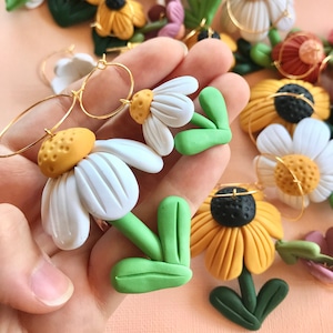 May include: A pair of handmade polymer clay earrings featuring white and yellow daisy flowers with green leaves. The earrings are attached to gold hoop earrings.