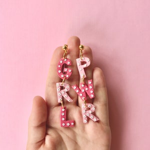 May include: Pink and white letter earrings spelling out "GIRL" with white polka dots. The earrings are hanging from a person's hand.