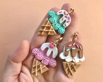 May include: Three pairs of ice cream cone earrings. Each pair features a different flavor of ice cream: mint, strawberry, and chocolate. The ice cream is topped with sprinkles and a cherry. The cones are made of a waffle pattern.