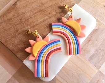 May include: A pair of rainbow and sun earrings made of polymer clay. The earrings are a bright pink, red, orange, yellow, blue, and green. The sun is a bright yellow with a pink outline.