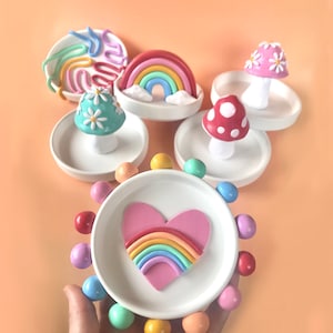 Puede incluir: Un conjunto de cinco platos de anillo de cer&aacute;mica coloridos con diferentes dise&ntilde;os. Cada plato presenta un arco&iacute;ris, un coraz&oacute;n con un arco&iacute;ris, un hongo con flores blancas o un dise&ntilde;o de espiral. Los platos son perfectos para guardar anillos, pendientes u otros art&iacute;culos peque&ntilde;os.