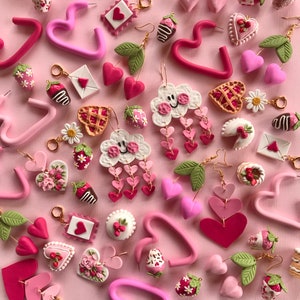 May include: Assortment of pink and red polymer clay earrings featuring various shapes, including hearts, clouds, strawberries, and flowers. The earrings are displayed on a pink background.