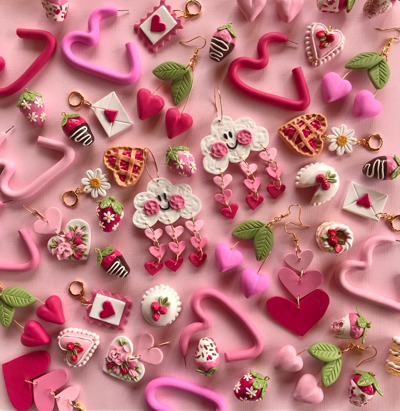 May include: Assortment of pink and red polymer clay earrings featuring various shapes, including hearts, clouds, strawberries, and flowers. The earrings are displayed on a pink background.