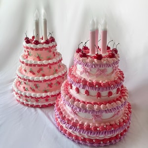 May include: Two pink and white tiered cakes decorated with cherries and white candles. The cakes are on a white surface.