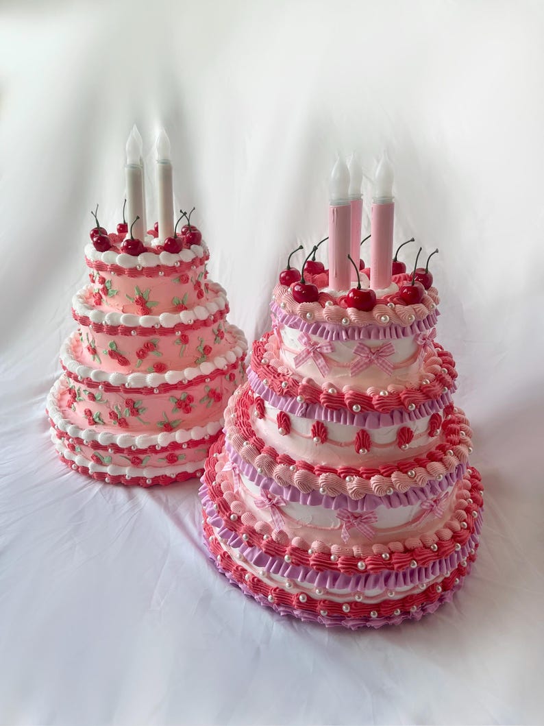 May include: Two pink and white tiered cakes decorated with cherries and white candles. The cakes are on a white surface.