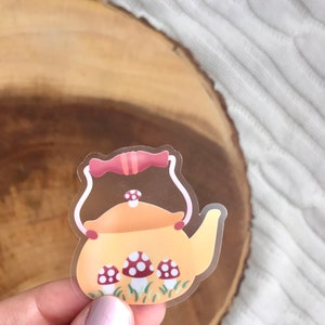 May include: A clear sticker with a yellow teapot with a red handle and lid. The teapot is surrounded by green grass and three red and white mushrooms.