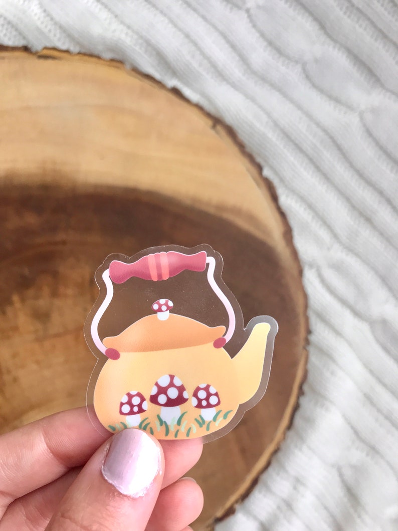May include: A clear sticker with a yellow teapot with a red handle and lid. The teapot is surrounded by green grass and three red and white mushrooms.