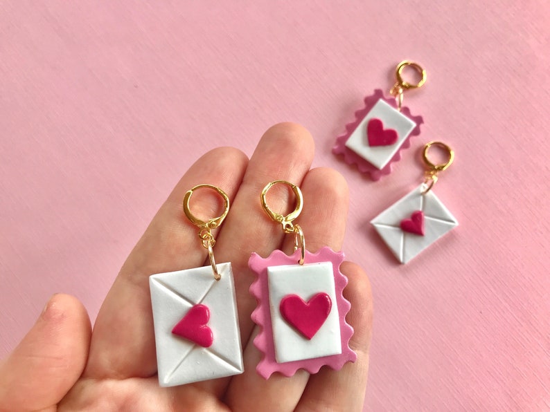 May include: Three pairs of pink and white polymer clay earrings shaped like envelopes with a pink heart on the front. The earrings are on a pink background.