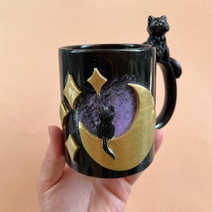 May include: Black ceramic mug with a gold crescent moon and stars design. A black cat figurine is perched on the handle.