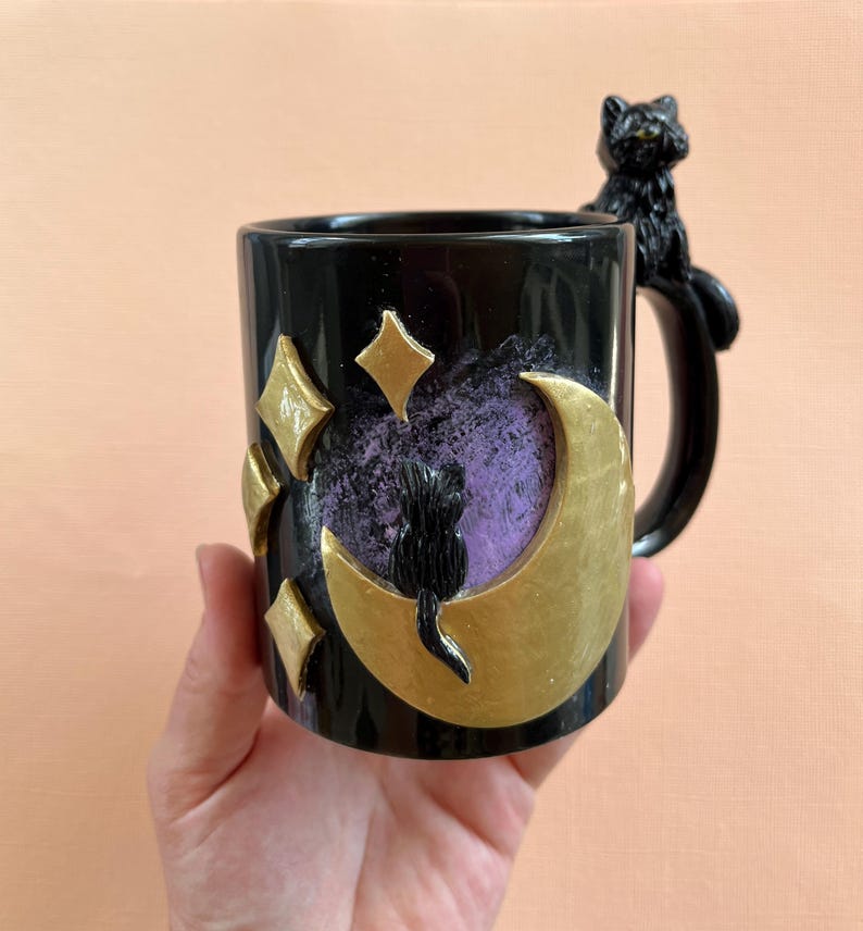 May include: Black ceramic mug with a gold crescent moon and stars design. A black cat figurine is perched on the handle.