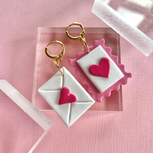 May include: A pair of earrings featuring a pink and white envelope design with a red heart on each. The earrings are made of polymer clay and have gold hoops.