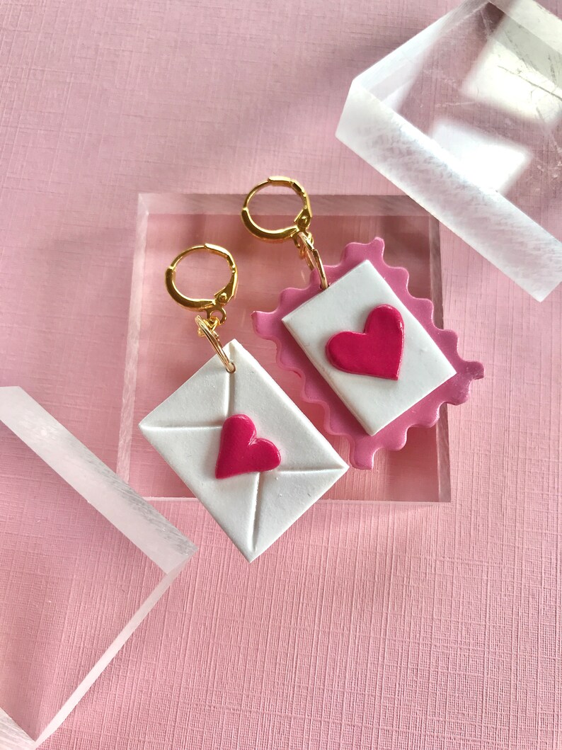 May include: A pair of earrings featuring a pink and white envelope design with a red heart on each. The earrings are made of polymer clay and have gold hoops.