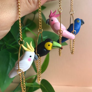 May include: Three colorful bird earrings with gold chains. The birds are a white cockatoo, a pink cockatoo, and a blue bird. The earrings are hanging from a gold chain.