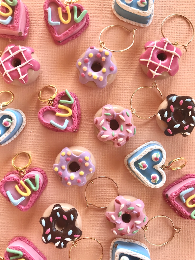 May include: A collection of colorful, handmade donut and heart-shaped charms with gold hoops. The donuts are decorated with sprinkles, icing, and chocolate. The hearts have the word "Luv" written on them.