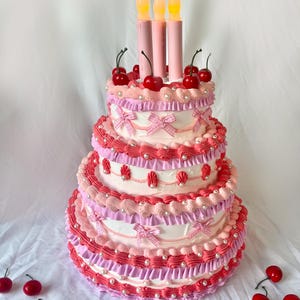 May include: A three-tiered pink and white cake decorated with cherries, bows, and pearl-like accents. The cake has three lit candles on top.