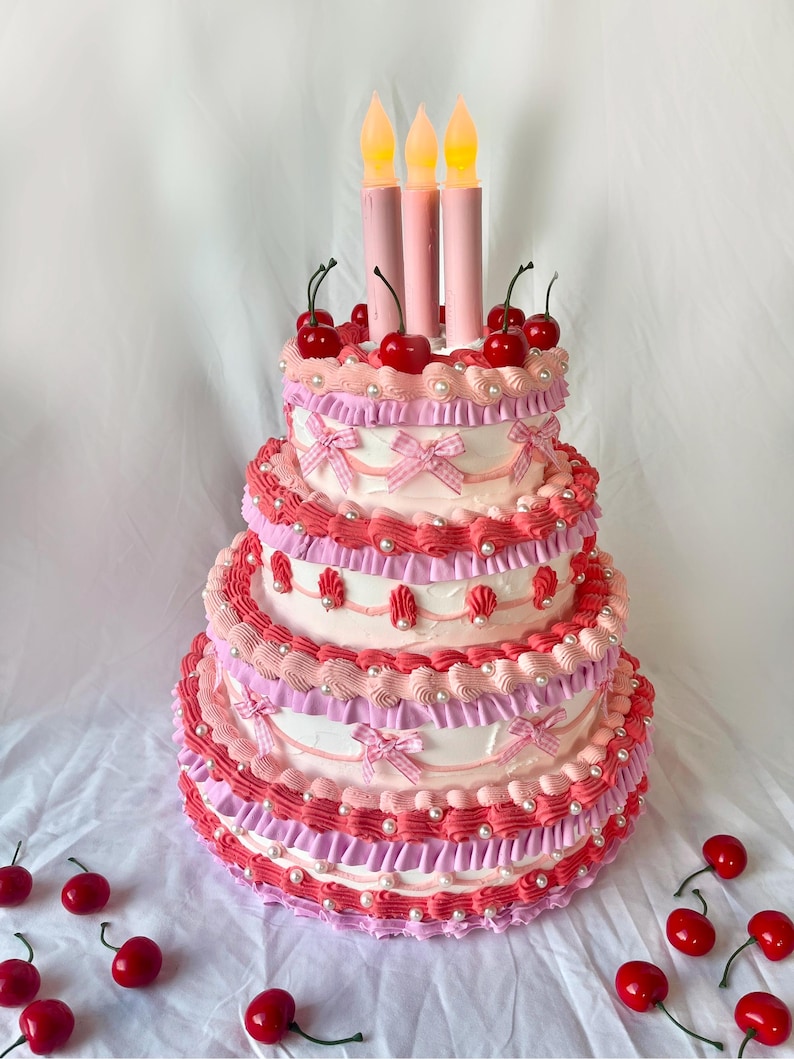 May include: A three-tiered pink and white cake decorated with cherries, bows, and pearl-like accents. The cake has three lit candles on top.