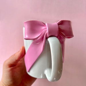 Pu&ograve; includere: Una tazza in ceramica bianca con un grande fiocco rosa attaccato al lato. Il fiocco &egrave; realizzato in un materiale morbido e flessibile e ha una finitura lucida.