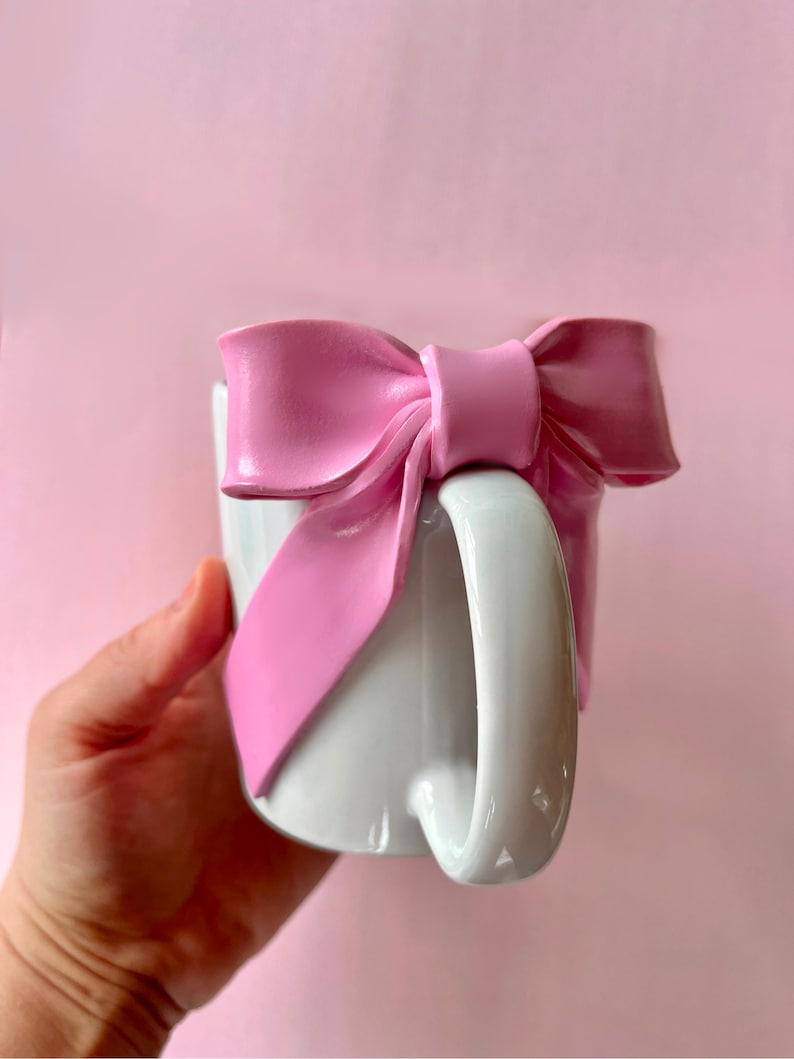 Pu&ograve; includere: Una tazza in ceramica bianca con un grande fiocco rosa attaccato al lato. Il fiocco &egrave; realizzato in un materiale morbido e flessibile e ha una finitura lucida.
