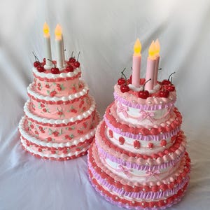 May include: Two tiered cakes decorated with pink and white frosting, cherries, and pearl accents. Both cakes have lit candles on top.