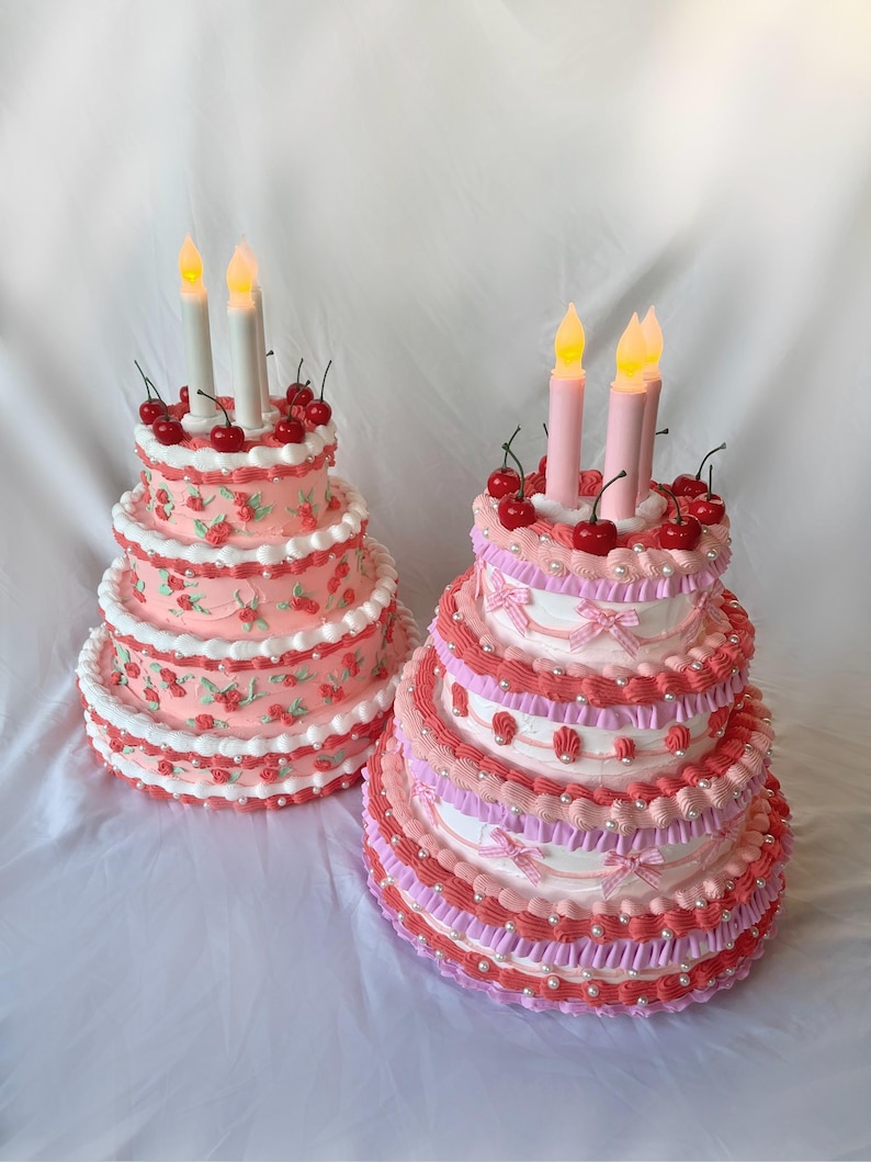 May include: Two tiered cakes decorated with pink and white frosting, cherries, and pearl accents. Both cakes have lit candles on top.
