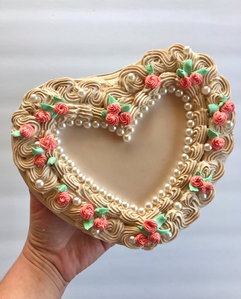 May include: A heart-shaped cake decorated with white frosting, pearl beads, and pink and green sugar flowers.