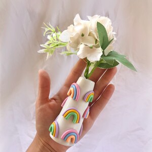 May include: White ceramic vase with a rainbow design. The vase is decorated with multiple pastel-colored rainbows. The vase is holding a bouquet of white flowers.