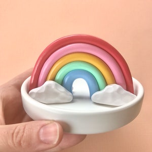 Puede incluir: Un peque&ntilde;o arco&iacute;ris hecho de arcilla de colores se sienta en un plato de cer&aacute;mica blanco con dos acentos en forma de nube blanca.