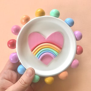 Puede incluir: Un plato de cer&aacute;mica blanco con un dise&ntilde;o de coraz&oacute;n arco&iacute;ris rodeado de bolas de colores.