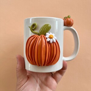 May include: White ceramic mug with a 3D orange pumpkin with a white flower and green stem. The pumpkin is on the front of the mug and the mug has a handle.