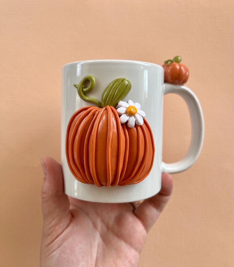 May include: White ceramic mug with a 3D orange pumpkin with a white flower and green stem. The pumpkin is on the front of the mug and the mug has a handle.