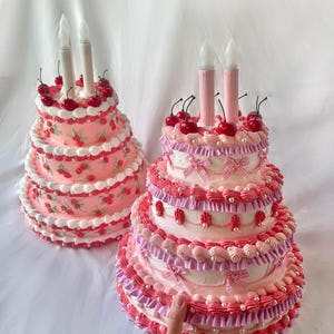 May include: Two pink and white tiered cakes decorated with cherries, white pearl beads, and pink and white ruffles. The cakes have white candles on top.