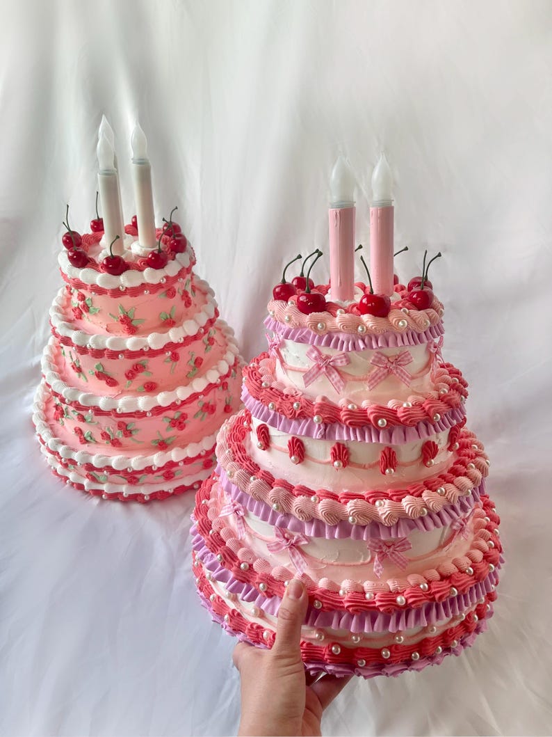 May include: Two pink and white tiered cakes decorated with cherries, white pearl beads, and pink and white ruffles. The cakes have white candles on top.
