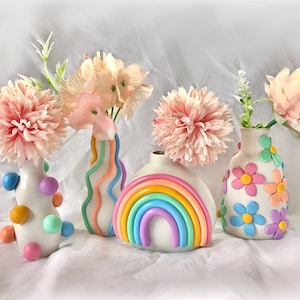 Puede incluir: Cuatro jarrones de cerámica con colores pastel y diseños diferentes. Los jarrones están decorados con rayas de arcoíris, lunares, flores y un arcoíris. Cada jarrón contiene un arreglo de flores rosas.