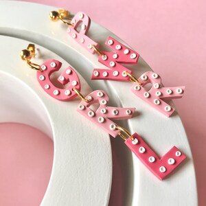 May include: Pink and white letter earrings spelling out "GIRL". Each letter is made of a pink and white ombre material with white dots.